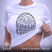 Футболка гори лікують. Біла футболка, складена на поверхні, фон світло-сірий, принт з горами та текстом чорного кольору, з етикеткою зеленого кольору.