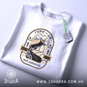 Футболка гори мій характер. Біла футболка, складена на поверхні, з принтом, що зображує гори та річку, виконаним у чорному та золотому кольорах. На футболці є етикетка з зеленим шнурком. Фон світло-фіолетовий.