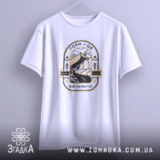 Футболка гори мій характер. Біла футболка, підвішена на вішалці, з принтом, що зображує гори та річку, виконаним у чорному та золотому кольорах. Фон світло-сірий, футболка має вільний крій.