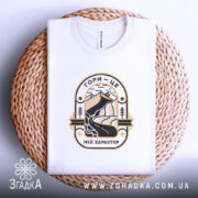 Футболка гори мій характер. Біла футболка, складена на плетеній кошику, з принтом, що зображує гори та річку, виконаним у чорному та золотому кольорах. Фон світло-фіолетовий, вид з верхнього ракурсу.