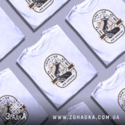 Футболка гори мій характер. Біла футболка з круглим вирізом, на моделі, з принтом, що зображує гори та річку, виконаним у чорному та золотому кольорах. Фон світло-фіолетовий, модель дивиться прямо в камеру.