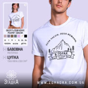 Футболка гори, біла, на моделі, з принтом гір та написом 'В ГОРАХ', фон світло-фіолетовий, деталі принта чорні, зображення чоловіка з коротким рукавом.