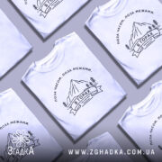 Футболка гори, біла, на моделі, з принтом гір та написом 'В ГОРАХ', фон світло-фіолетовий, деталі принта чорні, зображення чоловіка з коротким рукавом.