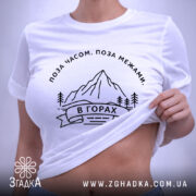 Футболка гори, біла, на плоскій поверхні, з принтом гір та написом 'В ГОРАХ', фон світло-фіолетовий, деталі принта чорні, з етикеткою з зеленим шнурком.