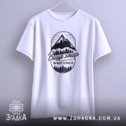 Футболка гори вище хмар, біла, підвішена на вішалці, фон світло-сірий, принт з горами та деревами чорного кольору з написом 'Вище хмар менше зайвого'.