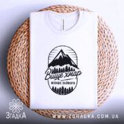 Футболка гори вище хмар, біла, складена на плетеній основі, фон світлий, принт з горами та деревами чорного кольору з написом 'Вище хмар менше зайвого'.