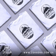 Футболка гори вище хмар, біла, зображена на моделі з легким ракурсом, фон ніжно-фіолетовий, принт з горами та деревами чорного кольору з написом 'Вище хмар менше зайвого'.