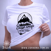 Футболка гори вище хмар, біла, складена на поверхні, з ракурсом зверху, фон світло-сірий, принт з горами та деревами чорного кольору з написом 'Вище хмар менше зайвого', прикріплена етикетка зеленого кольору.