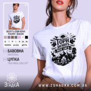 Футболка Гори знають, біла, на моделі з коротким рукавом, фон ніжно-фіолетовий, принт чорного кольору з гірським пейзажем, рюкзаком та слідами від черевиків.