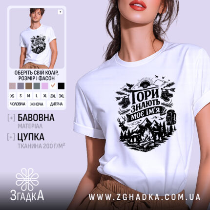 Футболка Гори знають, біла, на моделі з коротким рукавом, фон ніжно-фіолетовий, принт чорного кольору з гірським пейзажем, рюкзаком та слідами від черевиків.