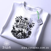 Футболка Гори знають, біла, лежить на плоскій поверхні, фон світлий, принт чорного кольору з гірським пейзажем, рюкзаком та слідами від черевиків, з зеленим ярликом.