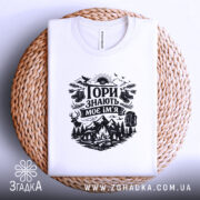 Футболка Гори знають, біла, лежить на плоскій поверхні, фон світлий, принт чорного кольору з гірським пейзажем, рюкзаком та слідами від черевиків, на плетеній основі.