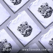 Футболка Гори знають, біла, на моделі з коротким рукавом, фон ніжно-фіолетовий, принт чорного кольору з гірським пейзажем, рюкзаком та слідами від черевиків.