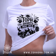 Футболка Гори знають, біла, лежить на плоскій поверхні, фон світлий, принт чорного кольору з гірським пейзажем, рюкзаком та слідами від черевиків, з зеленим ярликом.