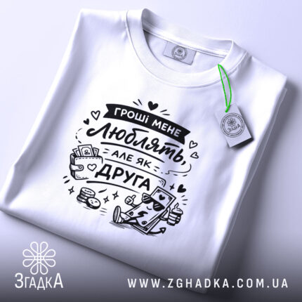 Футболка гроші мене люблять, біла, складена на поверхні, фон світло-сірий, принт чорного кольору з малюнком грошей і написом, з зеленим ярликом.