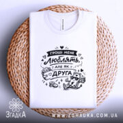 Футболка гроші мене люблять, біла, складена на плетеній кошику, фон світло-сірий, принт чорного кольору з малюнком грошей і написом.