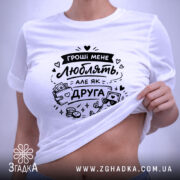 Футболка гроші мене люблять, біла, складена на поверхні, фон світло-сірий, принт чорного кольору з малюнком грошей і написом, з зеленим ярликом.