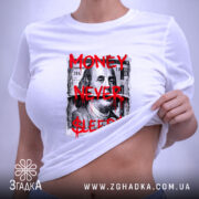 Футболка Гроші не сплять, біла, складена на поверхні, ракурс зверху, фон світло-сірий, принт з написом 'MONEY NEVER $LEEPS$' червоного кольору на фоні зображення купюри, з етикеткою зеленого кольору.