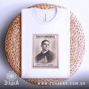 Футболка Григорій Сковорода, біла, зображена на столі, ракурс зверху, фон світло-сірий, принт у вигляді поштової марки з портретом Григорія Сковороди та написом 'SVIT LOVYV MENE', футболка лежить на плетеній основі.