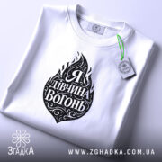 Футболка я дівчина вогонь, біла, на плоскій поверхні, з принтом у вигляді вогню та написом "Я Дивчина Вогонь", етикетка з зеленим шнурком, фон світлий.