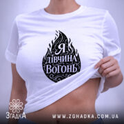 Футболка я дівчина вогонь, біла, на плоскій поверхні, з принтом у вигляді вогню та написом "Я Дивчина Вогонь", етикетка з зеленим шнурком, фон світлий.