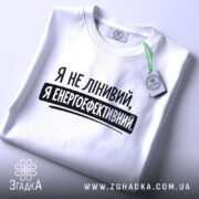 Футболка я енергоефективний, біла, складена на поверхні, ракурс зверху, фон світло-сірий, принт чорного кольору з текстом 'Я не лінивий, я енергоефективний', з етикеткою зеленого кольору.
