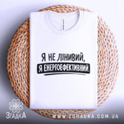 Футболка я енергоефективний, біла, складена на плетеній основі, ракурс зверху, фон світло-сірий, принт чорного кольору з текстом 'Я не лінивий, я енергоефективний'.