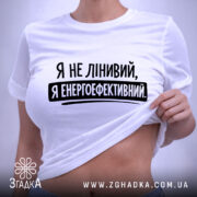 Футболка я енергоефективний, біла, складена на поверхні, ракурс зверху, фон світло-сірий, принт чорного кольору з текстом 'Я не лінивий, я енергоефективний', з етикеткою зеленого кольору.