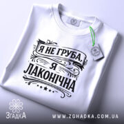 Футболка я не груба. Біла футболка, складена на плоскій поверхні, зображена зверху. Принт чорного кольору з написом "Я не груба, я лаконічна" займає центральну частину. На футболці присутня етикетка з зеленим шнурком.