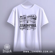 Футболка я не груба. Біла футболка з коротким рукавом, підвішена на вішалці, зображена з фронтального ракурсу. Принт чорного кольору з написом "Я не груба, я лаконічна" розташований по центру, фон світло-сірий.