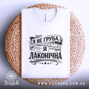 Футболка я не груба. Біла футболка, складена на круглому плетеному підносі, зображена зверху. Принт чорного кольору з написом "Я не груба, я лаконічна" займає центральну частину, фон білий.