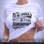 Футболка я не груба. Біла футболка, складена на плоскій поверхні, зображена зверху. Принт чорного кольору з написом "Я не груба, я лаконічна" займає центральну частину. На футболці присутня етикетка з зеленим шнурком.