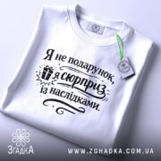 Футболка я не подарунок, біла футболка, складена на поверхні, з принтом чорного кольору, текст "Я не подарунок, я сюрприз із наслідками" та зображенням подарунка, на фоні світло-сірого кольору.
