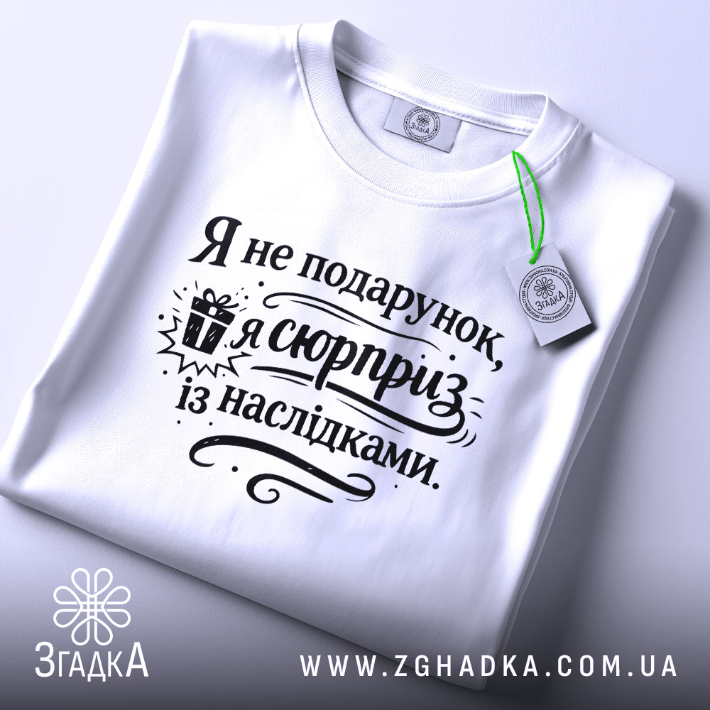 Футболка я не подарунок, біла футболка, складена на поверхні, з принтом чорного кольору, текст "Я не подарунок, я сюрприз із наслідками" та зображенням подарунка, на фоні світло-сірого кольору.