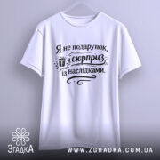 Футболка я не подарунок, біла футболка, підвішена на вішалці, з коротким рукавом, принт чорного кольору з текстом "Я не подарунок, я сюрприз із наслідками", на фоні світло-сірого кольору.