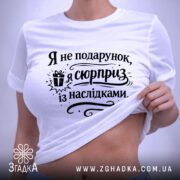 Футболка я не подарунок, біла футболка, складена на поверхні, з принтом чорного кольору, текст "Я не подарунок, я сюрприз із наслідками" та зображенням подарунка, на фоні світло-сірого кольору.