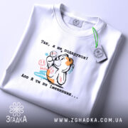 Футболка Я не подарунок. Біла футболка, складена на столі, фон світло-сірий. Принт з ведмедиком, який тримає кульки, з написом українською мовою, видна етикетка з зеленим шнурком.