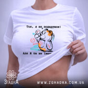 Футболка Я не подарунок. Біла футболка, складена на столі, фон світло-сірий. Принт з ведмедиком, який тримає кульки, з написом українською мовою, видна етикетка з зеленим шнурком.