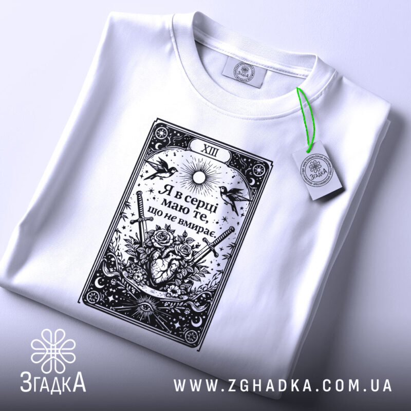 Футболка Я в серці має білий колір, зображена в горизонтальному ракурсі на плоскій поверхні. Принт чорного кольору з елементами графіки, що включає серце, квіти та мечі, розташований по центру. На футболці видно етикетку з зеленим шнурком, фон світлий.