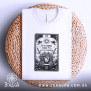 Футболка Я в серці має білий колір, зображена на плоскій поверхні, складена на круглій кошику з натурального матеріалу. Принт чорного кольору з елементами графіки, що включає серце, квіти та мечі, розташований по центру. Фон світлий, що підкреслює текстуру тканини.