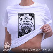 Футболка Я в серці має білий колір, зображена в горизонтальному ракурсі на плоскій поверхні. Принт чорного кольору з елементами графіки, що включає серце, квіти та мечі, розташований по центру. На футболці видно етикетку з зеленим шнурком, фон світлий.