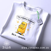 Футболка я важка людина. Біла футболка, складена на поверхні, з принтом жовтого злитка золота. Принт має яскраві кольори, текст зверху: 'Так, я важка людина!' Фон світлий, акцент на футболці.