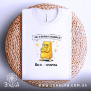 Футболка я важка людина. Біла футболка, складена на плетеній кошику, з принтом жовтого злитка золота. Принт яскравий, текст зверху: 'Так, я важка людина!' Фон світлий, акцент на футболці.