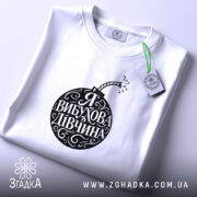 Футболка я вибухова дівчина, біла, зображена зверху на плоскості, фон світло-фіолетовий, принт чорного кольору у формі бомби з написом 'Я ВИБУХОВА ДІВЧИНА', з етикеткою зеленого кольору.