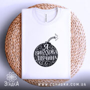 Футболка я вибухова дівчина, біла, зображена зверху на плоскості, фон світло-фіолетовий, принт чорного кольору у формі бомби з написом 'Я ВИБУХОВА ДІВЧИНА', на плетеній основі.