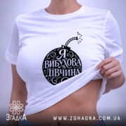 Футболка я вибухова дівчина, біла, зображена зверху на плоскості, фон світло-фіолетовий, принт чорного кольору у формі бомби з написом 'Я ВИБУХОВА ДІВЧИНА', з етикеткою зеленого кольору.