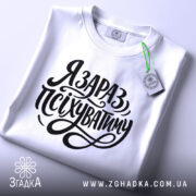 Футболка я зараз псіхуватиму, біла, на плоскій поверхні, фон світлий, принт чорний, з написом 'Я ЗАРАЗ, ПСІХУВАТИМУ' у стилі рукописного шрифту, з етикеткою на комірці.