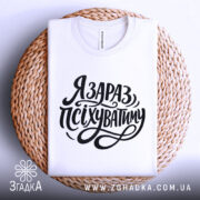 Футболка я зараз псіхуватиму, біла, складена на плоскій поверхні, фон світлий, принт чорний, з написом 'Я ЗАРАЗ, ПСІХУВАТИМУ' у стилі рукописного шрифту, на плетеній основі.