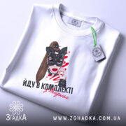 Футболка йду в комплекті з валідолом, біла, зображена на плоскій поверхні, фон світло-фіолетовий, принт з жінкою в масці кота, написом і етикеткою з зеленим шнурком.