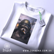 Футболка єнот пілот, біла, на плоскій поверхні, з верхнього ракурсу, фон світлий, принт з єнотом у пілотському шоломі з сигаретою, на фоні літаки та вибухи, з етикеткою.